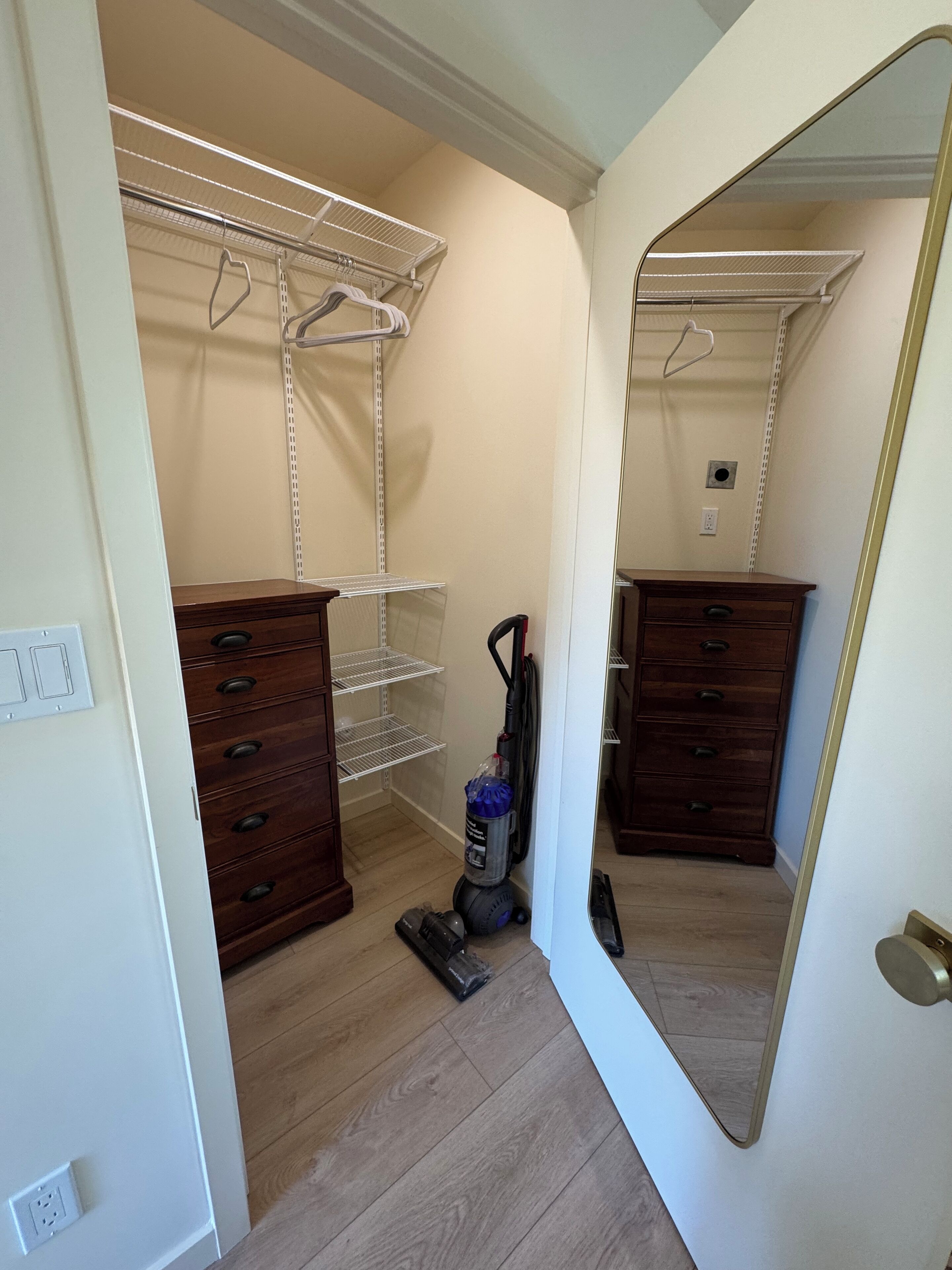 A walk-in closet with wooden dressers, metal shelving units, and a vacuum cleaner.