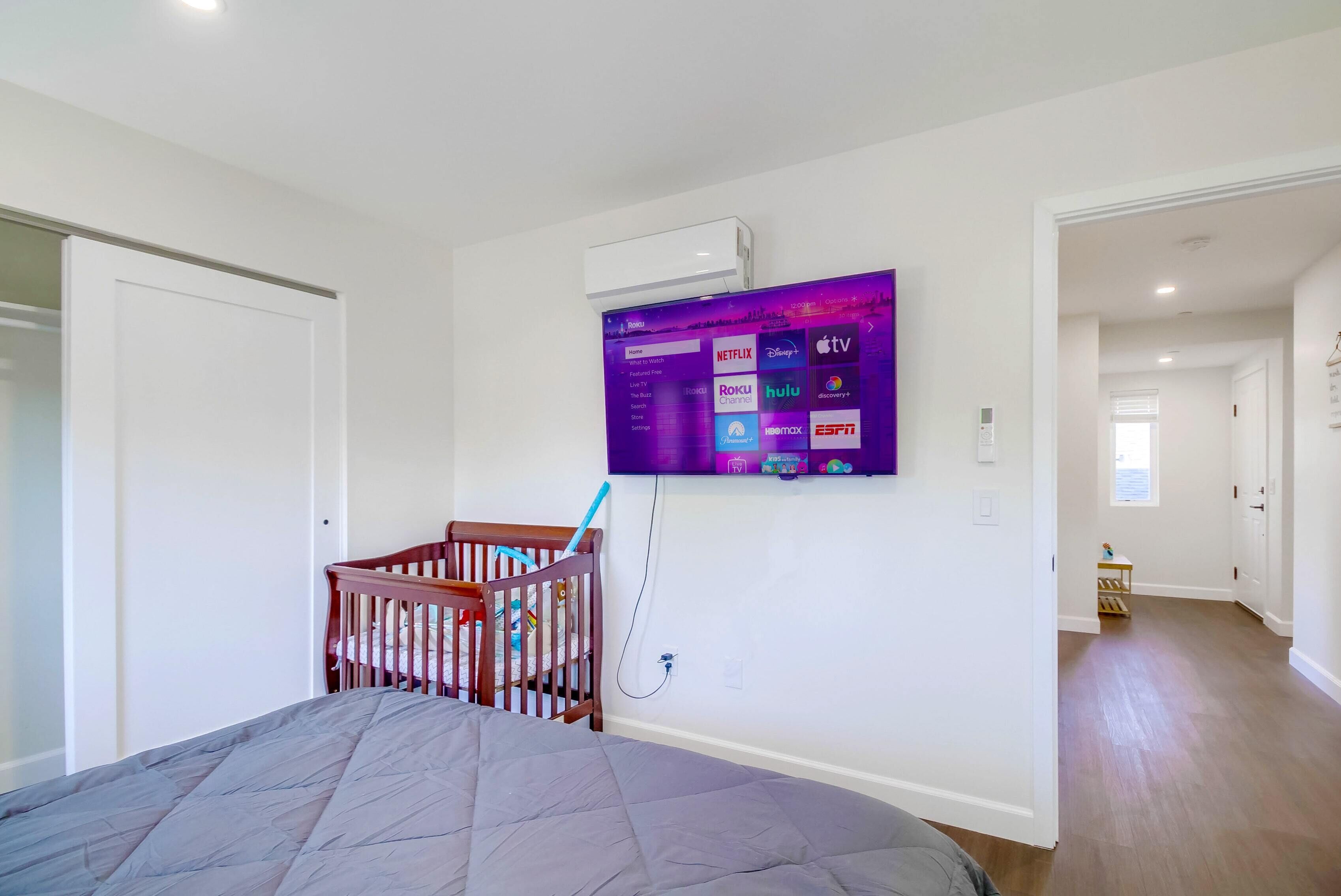 A bedroom with a bed, a crib, a mounted TV displaying streaming services, and a hallway leading to another room.