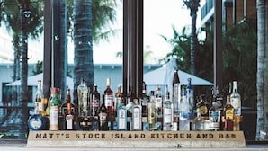 Baaritiski, jonka päällä on monenlaisia alkoholijuomapulloja, ja sen yläpuolella on kyltti, jossa lukee: ”MATT’S STOCK ISLAND KITCHEN AND BAR”.