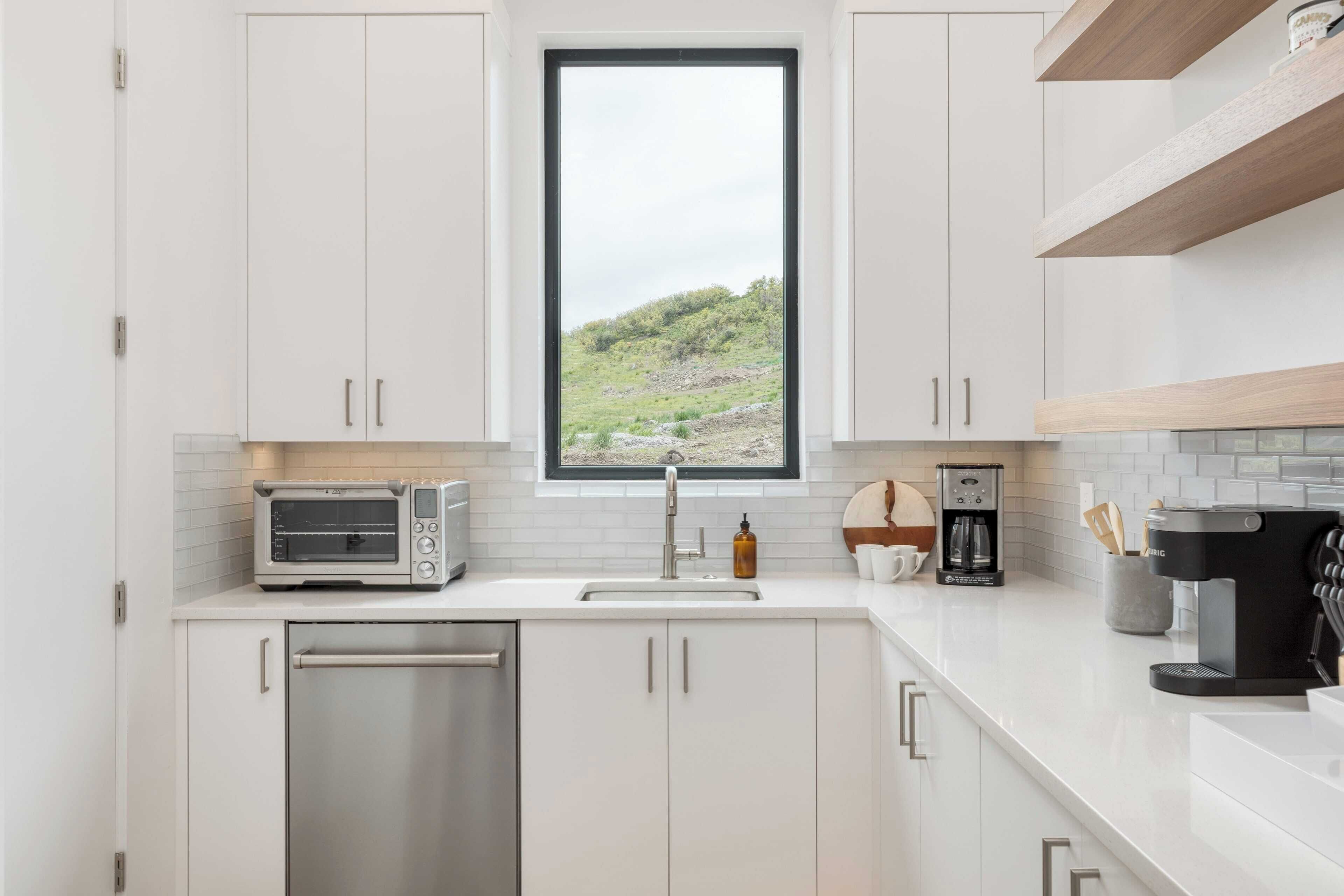 A modern kitchen with white cabinets, a built-in oven, a toaster, a coffee maker, and a window with a view of a green landscape.