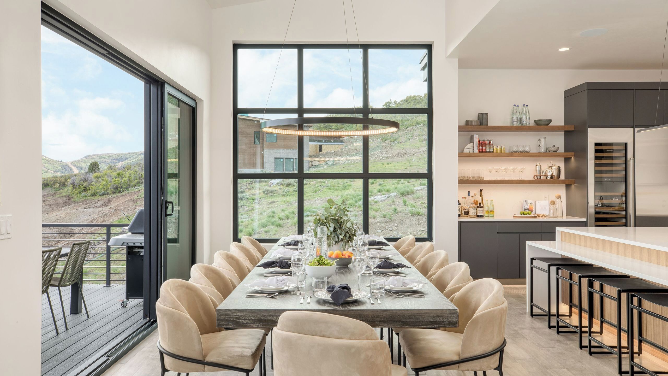Un espace de repas moderne avec une grande table dressée pour six personnes, une cuisine aux meubles sombres et une vue sur un paysage vallonné grâce à de grandes fenêtres.