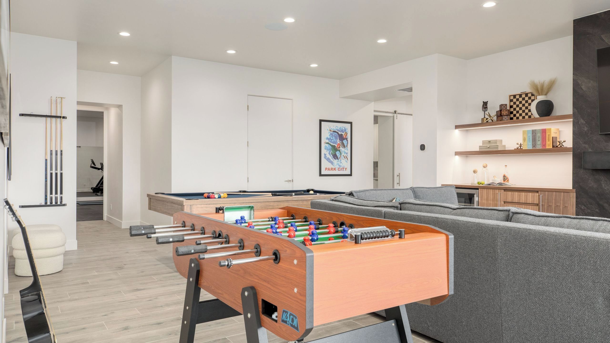 A modern living room with a foosball table, grey sofa, and a bookshelf with books and decorative items.