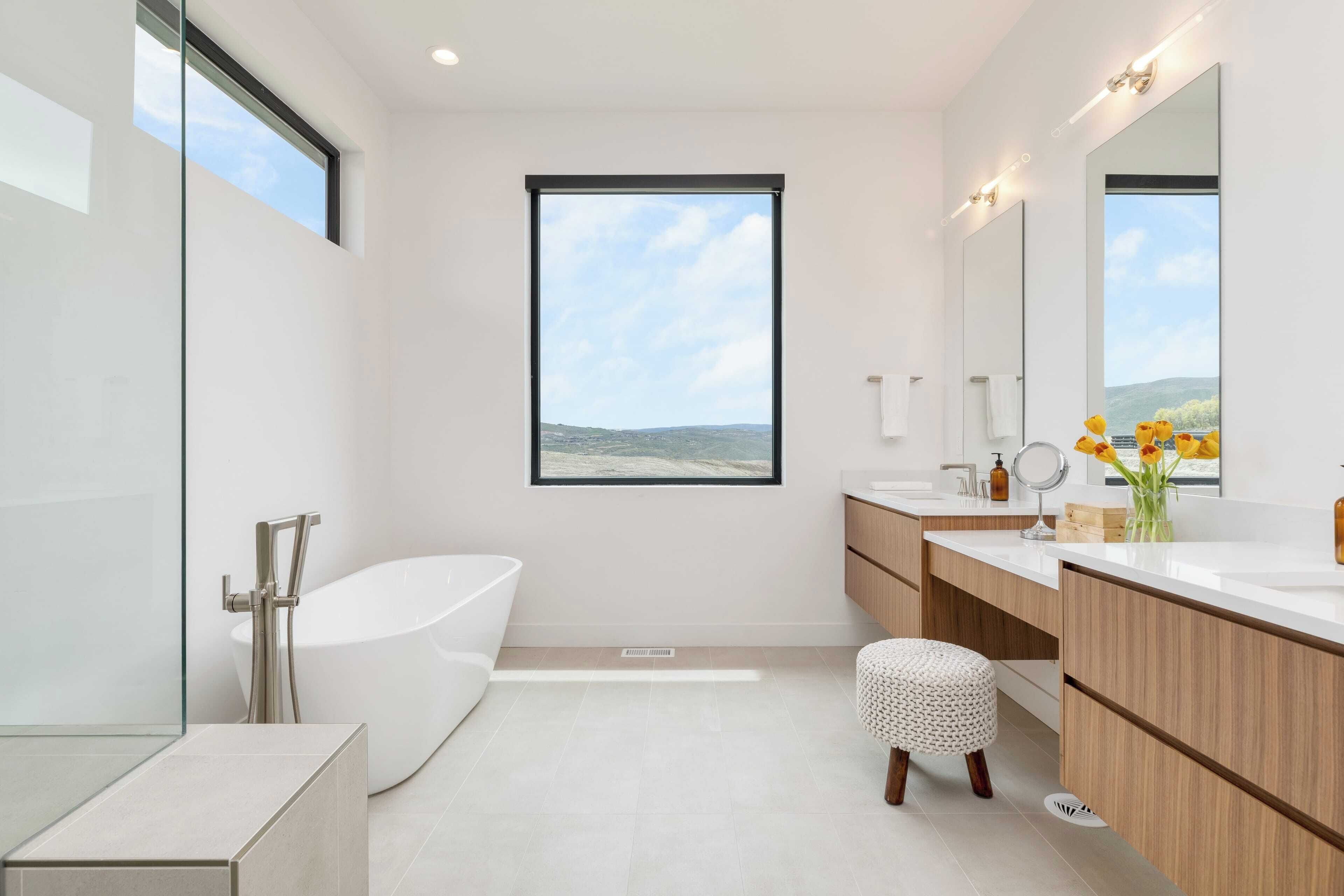 A modern bathroom with a large window, a freestanding bathtub, a vanity with a mirror, and a small stool.