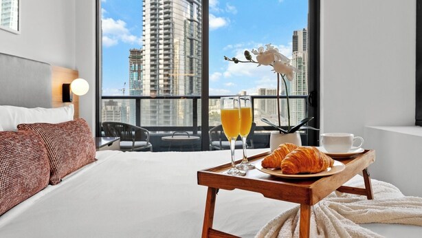 A hotel room with a large window, a bed, a breakfast tray with croissants, juice, and a cup, and a view of tall buildings. - NEW 2BD/2BR  WorldCenter Miami - Amazing views (Miami)
