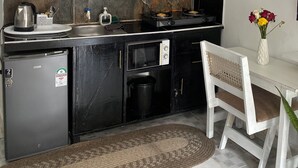 Een compacte keuken met een koelkast, magnetron, fornuis en een klein spoelgedeelte. Er is een stoel en een tafel met een bloevas.