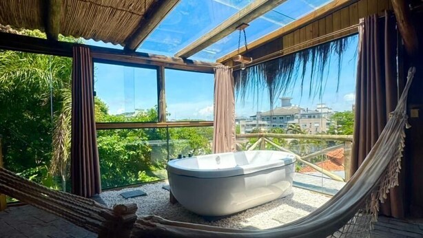 A hammock in a room with a glass ceiling, a bathtub, and a view of greenery and buildings. - Pousada Asa Delta Jurerê (Florianopolis)