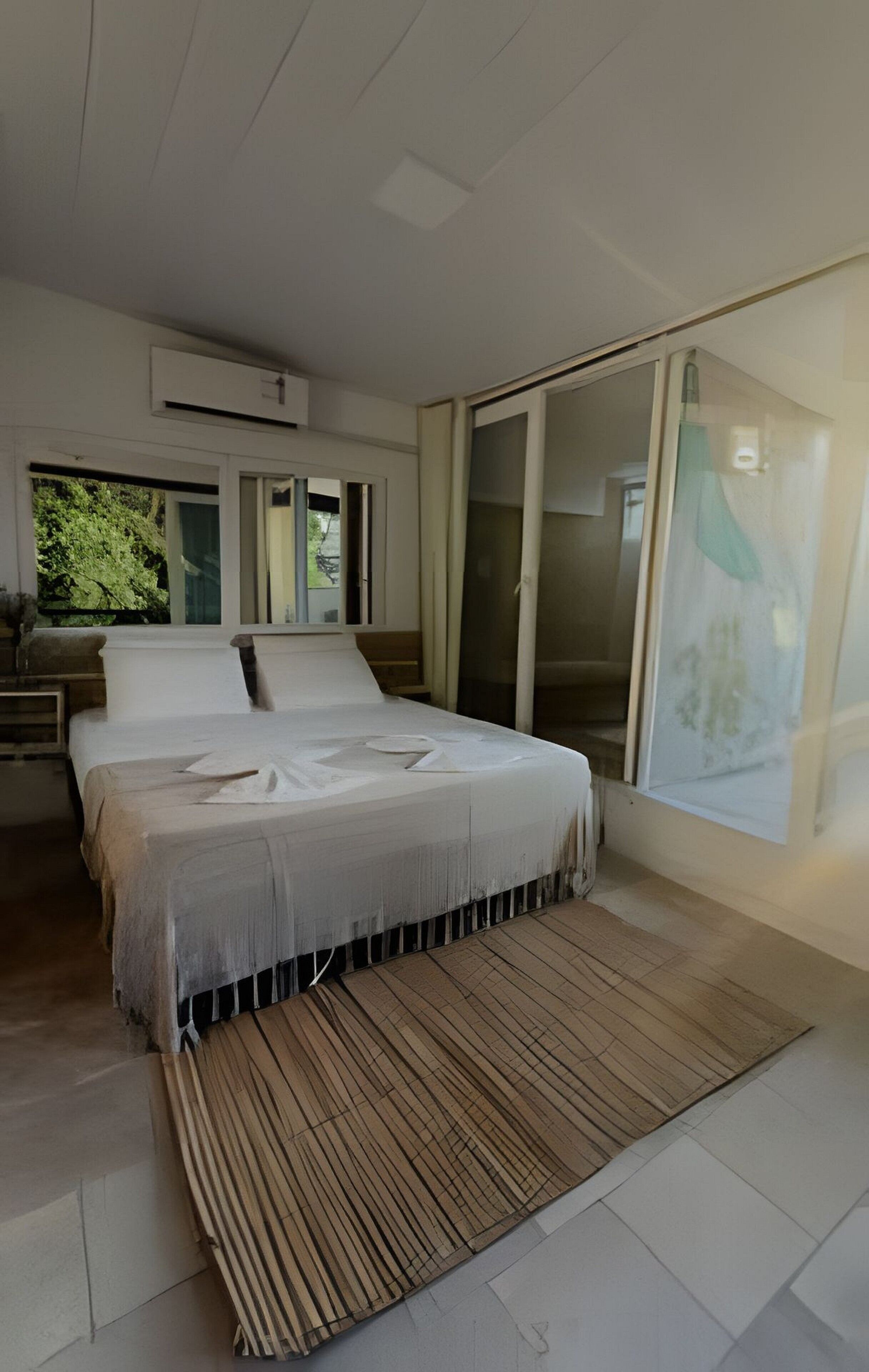 A modern bedroom with a large bed, a wooden floor, and a view of greenery through the window.