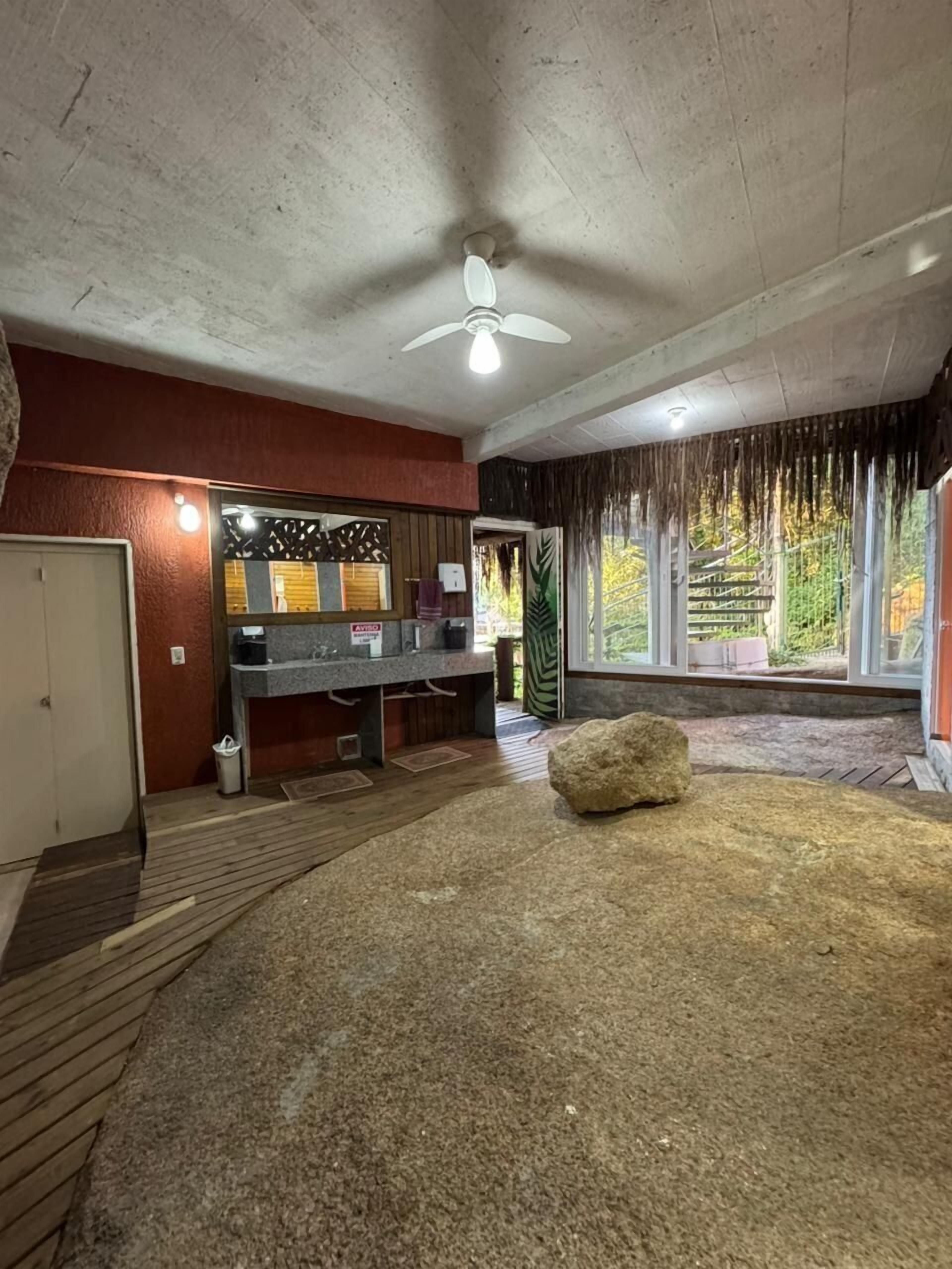 A room with a wooden floor, a large rock, a ceiling fan, and a window with a view of greenery.