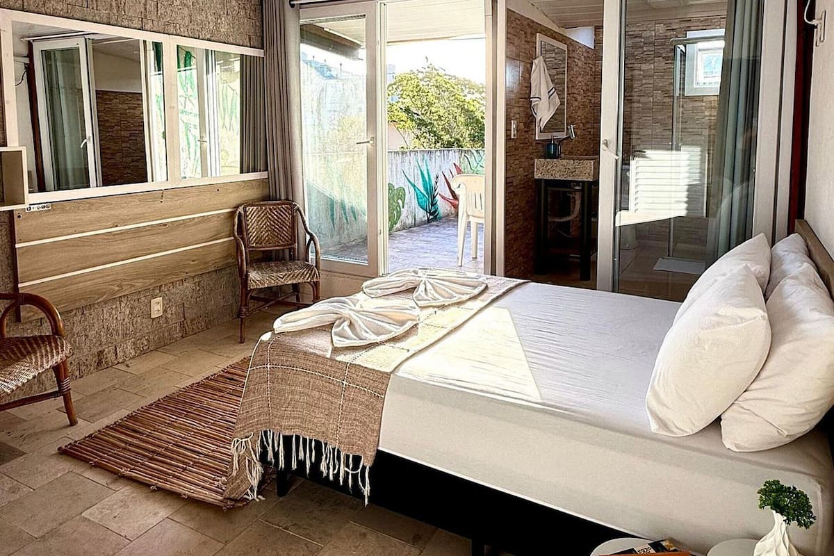 A bedroom with a bed, a small table, a chair, a window, and a bathroom with a shower.