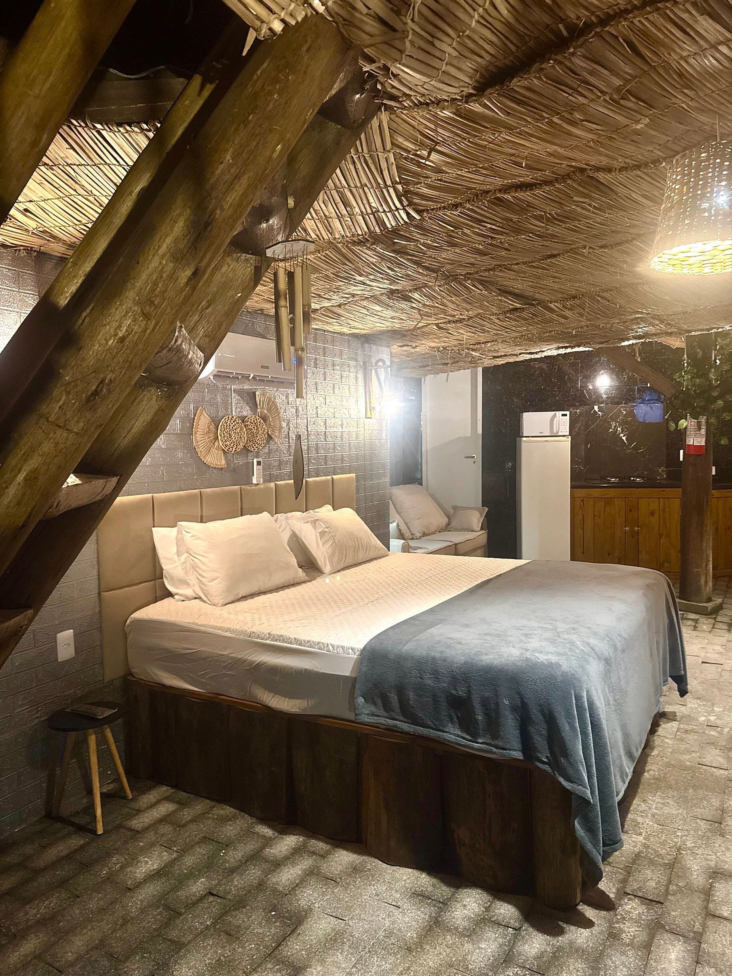 A cozy room with a bed, a small table, and a chair. There is a wooden ceiling and a brick wall.
