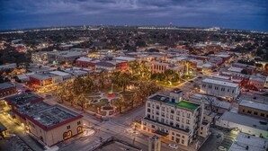 Une vue aérienne d’une ville au crépuscule, avec des bâtiments illuminés, une place centrale bordée d’arbres et un clocher d’église imposant.