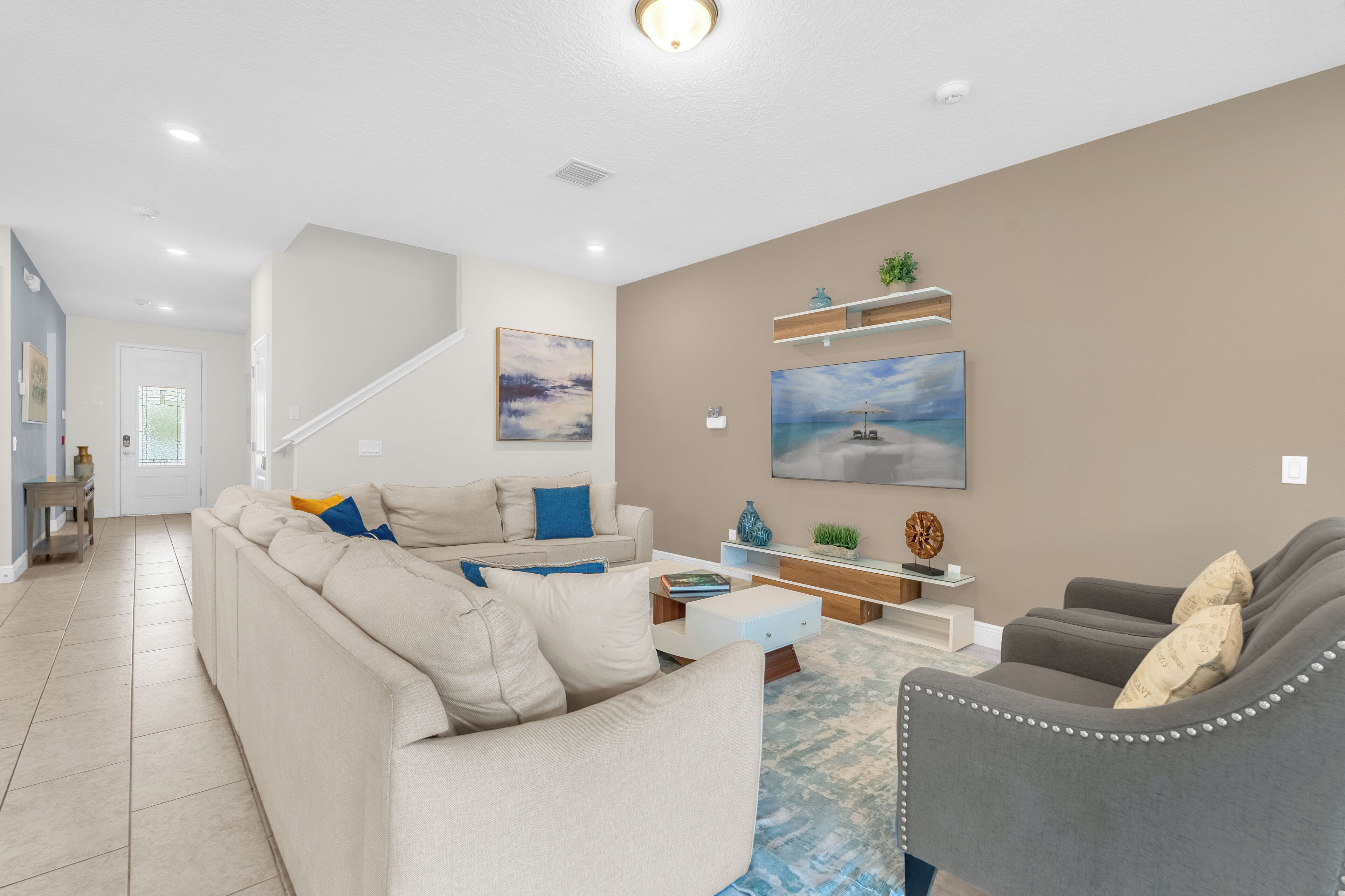 A modern living room with a beige sofa, a grey armchair, and a coffee table. There is a painting on the wall and a staircase in the background.