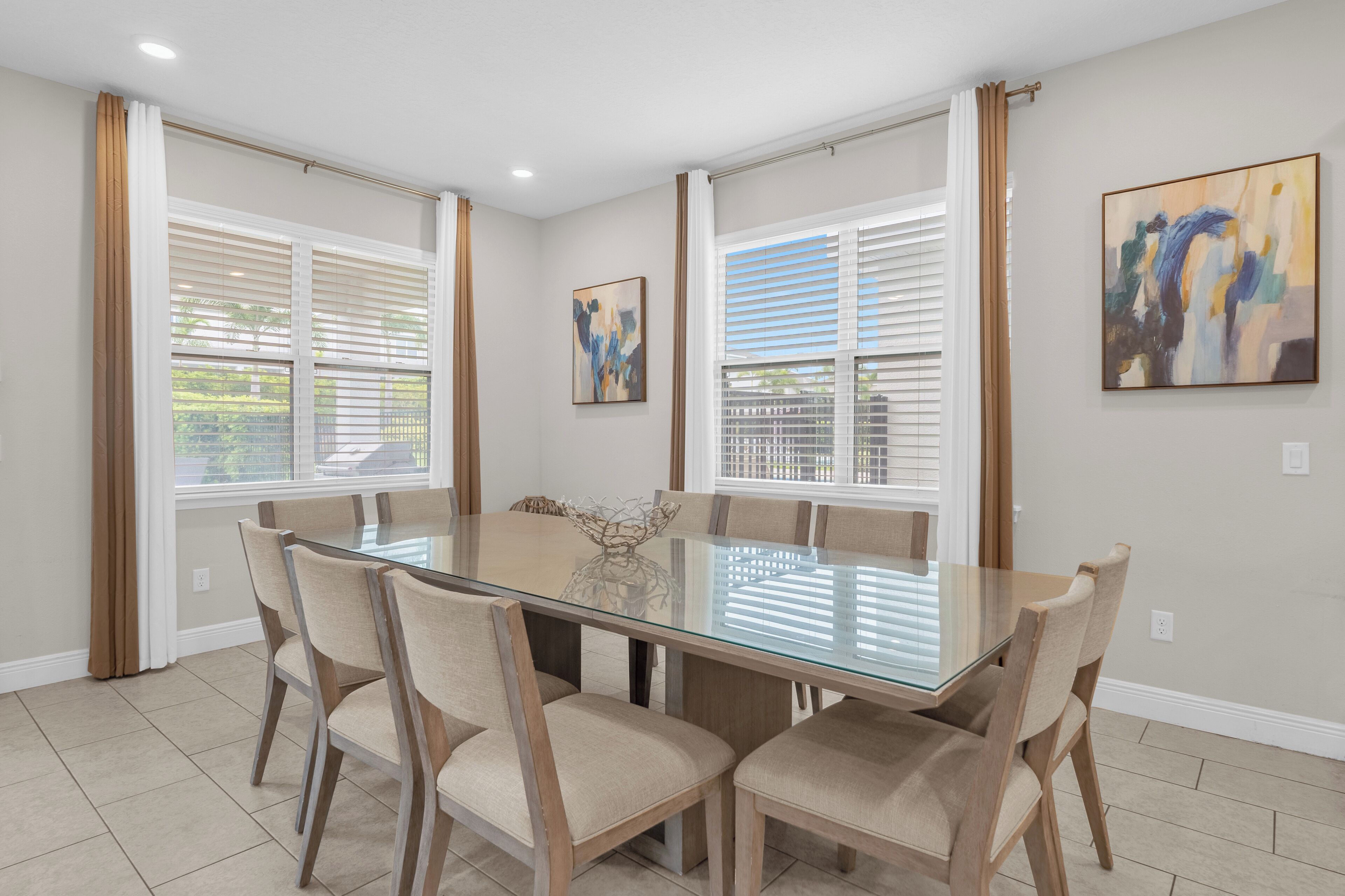 Une salle à manger avec une table à plateau de verre entourée de six chaises, de grandes fenêtres équipées de stores et deux œuvres d’art abstraites accrochées aux murs.