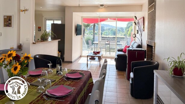 Un comedor con una mesa preparada para cuatro personas, un ventilador de techo y vistas al exterior a través de un gran ventanal corredero.