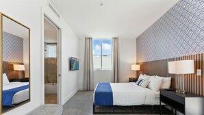 A modern hotel room with a large bed, bedside tables, a bench, a shower area, and a view of the sky through the window. - South Beach Sleek 2BR Getaway (Miami Beach)