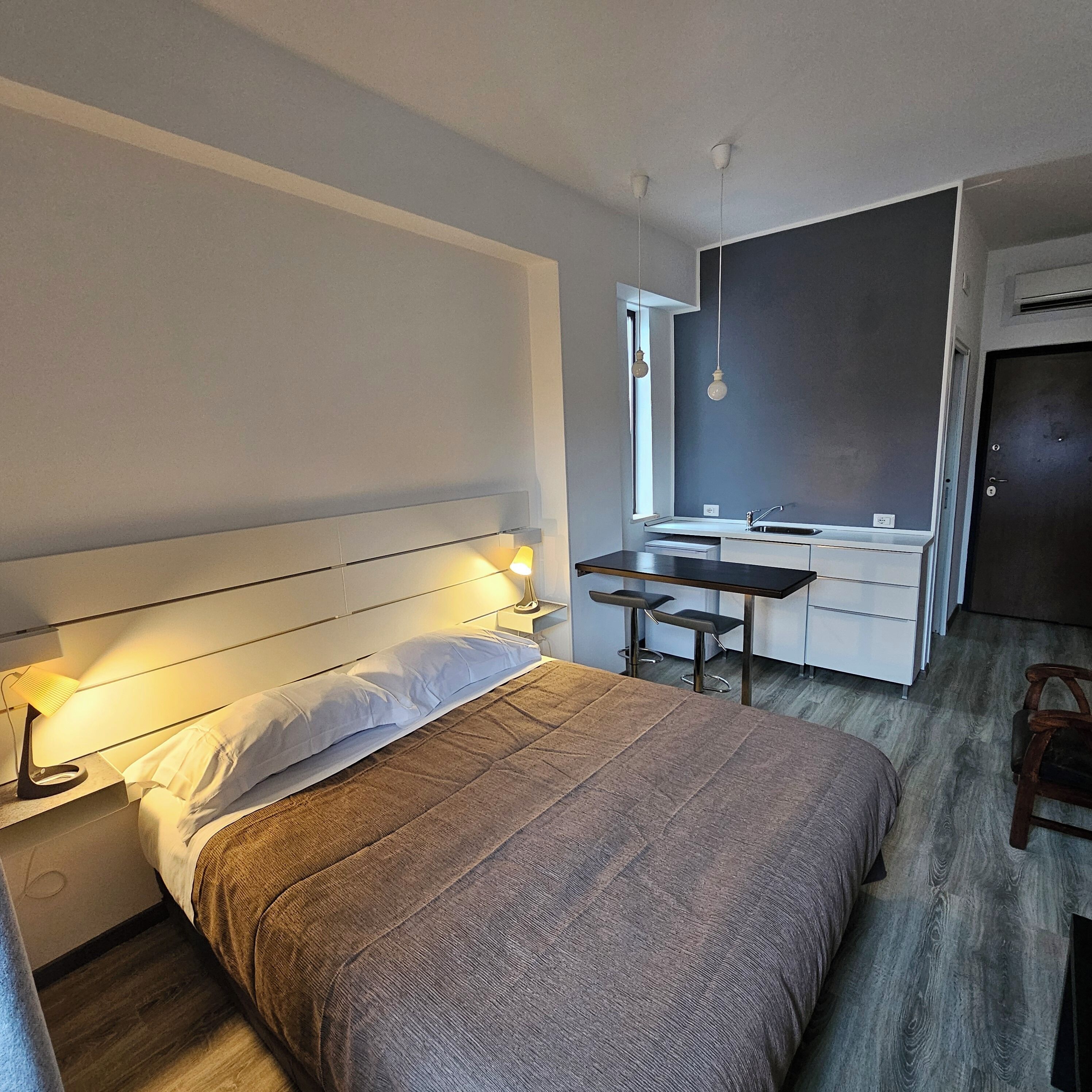 Ein Schlafzimmer mit Bett, Nachttischlampe und einer kleinen Küchenzeile mit Spüle und Barhocker.