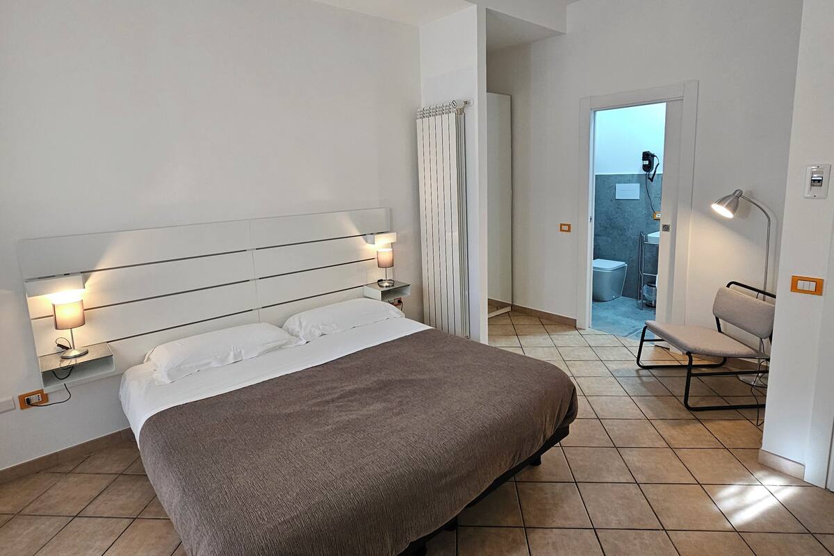 Ein modernes Schlafzimmer mit einem großen Bett, Nachttischlampen, einer Heizung und Blick in ein Badezimmer durch eine offene Tür.