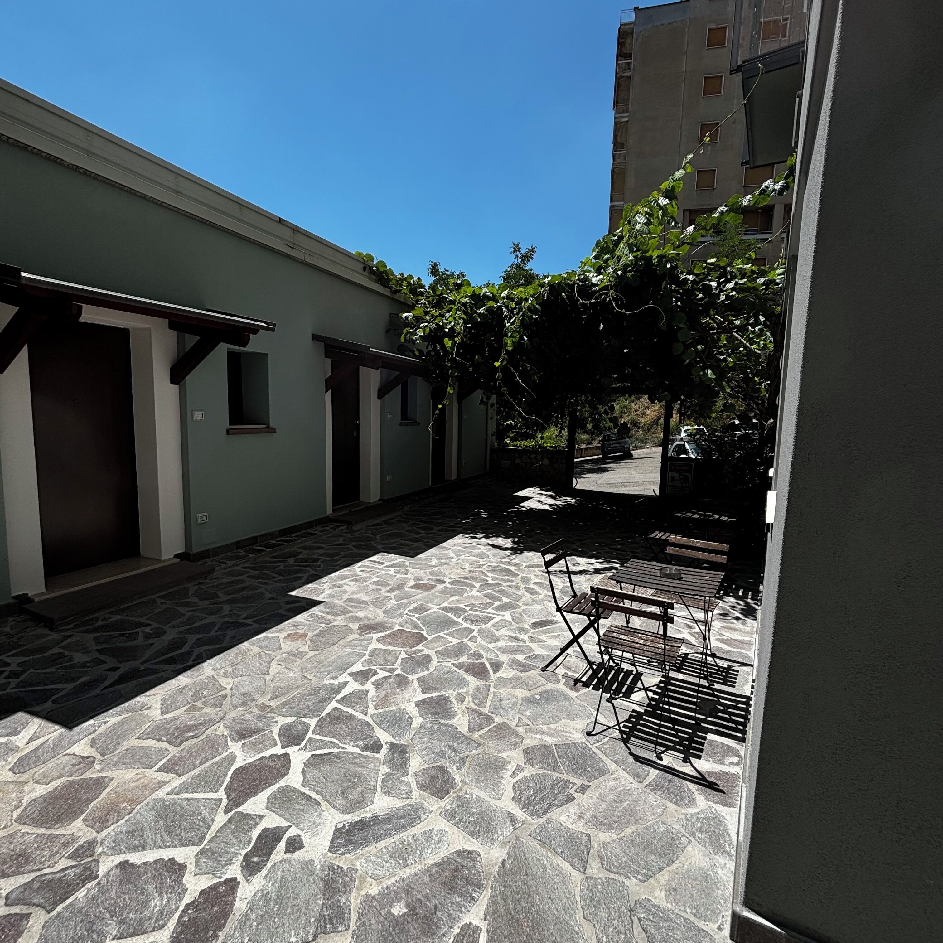 Eine Terrasse im Freien mit Steinboden, einer Pergola mit Kletterpflanzen und einer Sitzecke mit Metallstühlen und -tischen.