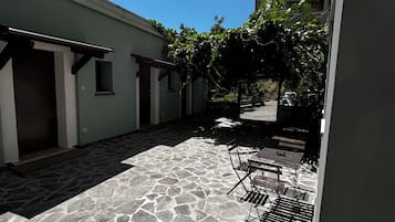 Eine Terrasse im Freien mit Steinboden, einer Pergola mit Kletterpflanzen und einer Sitzecke mit Metallstühlen und -tischen.