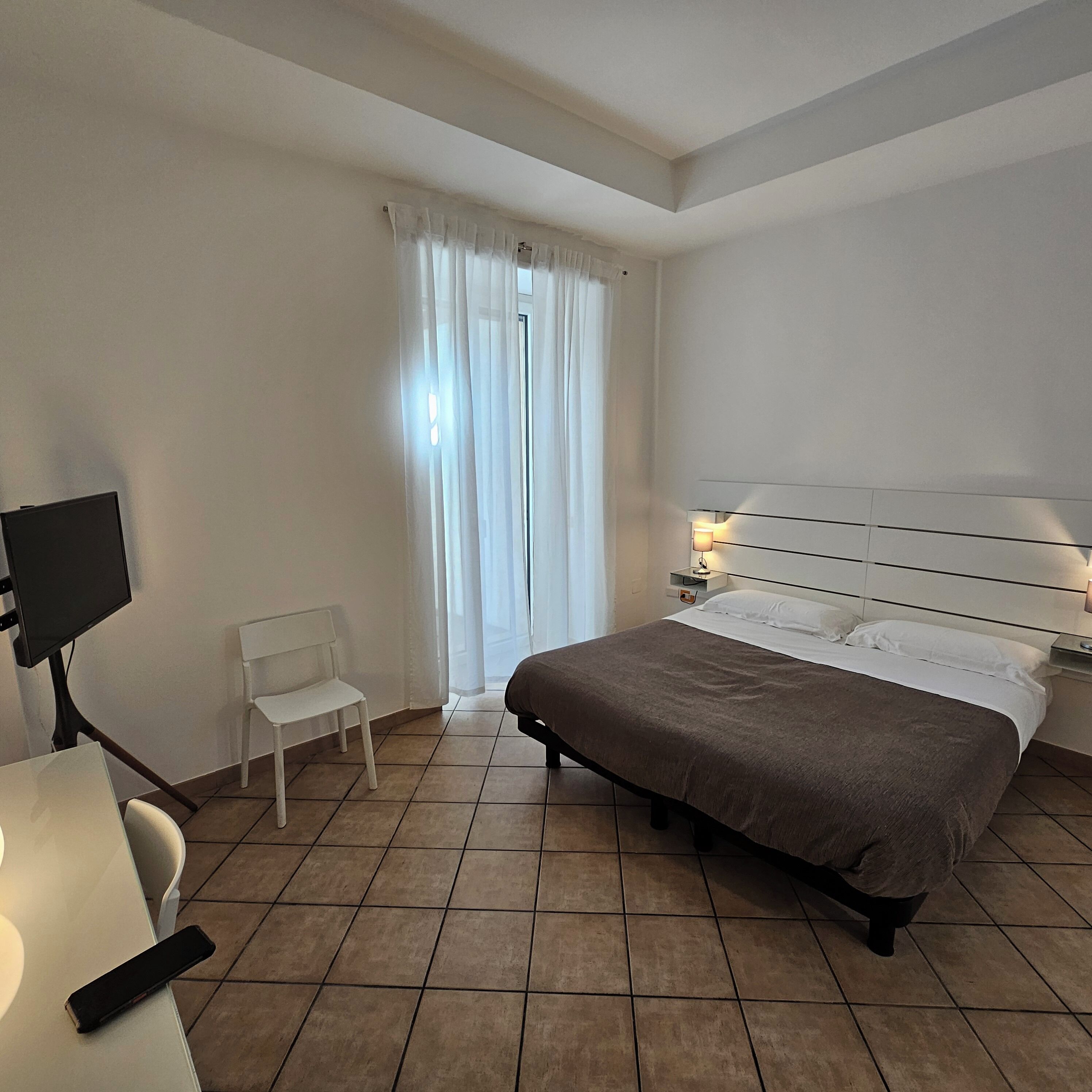 A hotel room with a bed, a chair, a desk with a computer monitor, and a nightstand with a lamp.