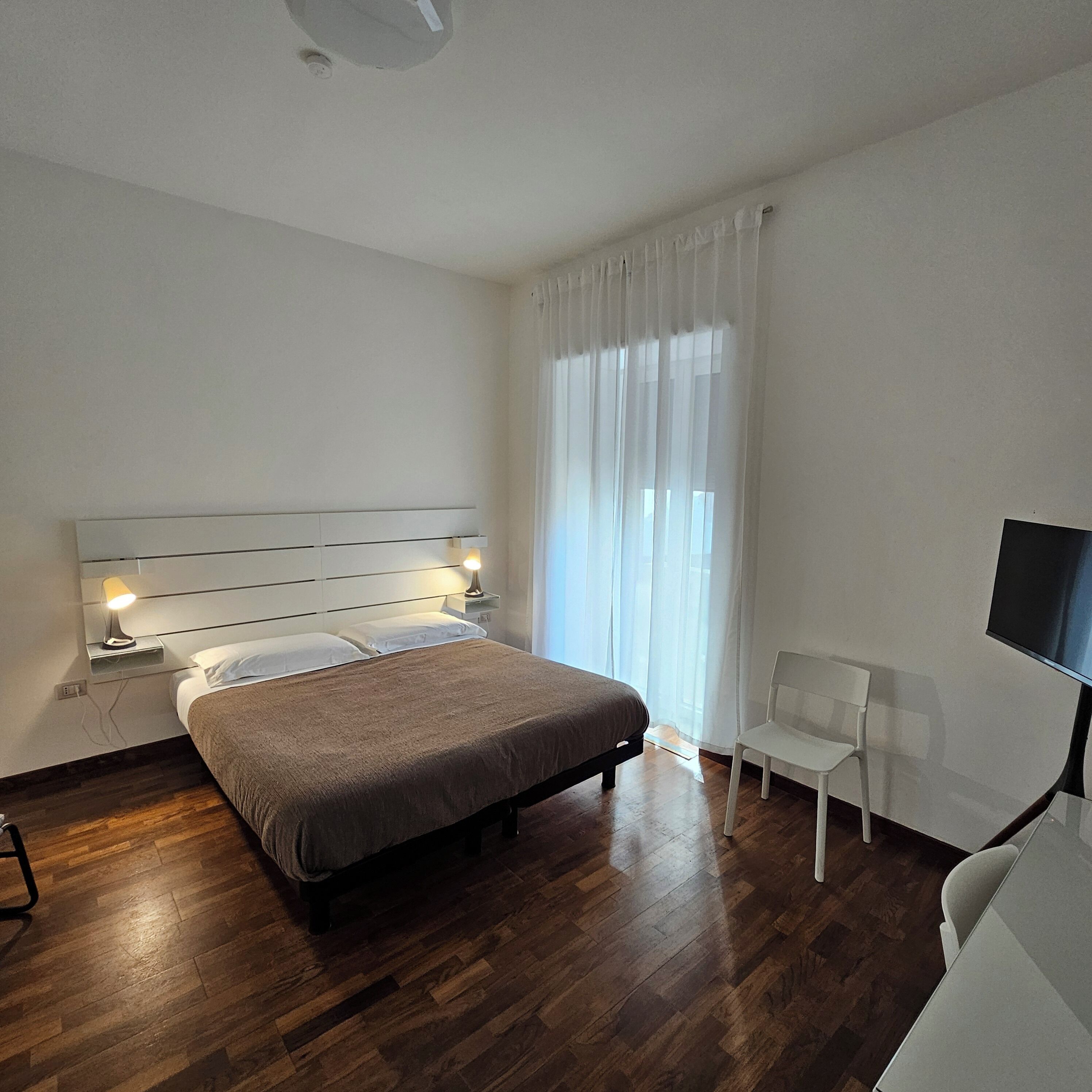 A modern bedroom with a wooden floor, a bed with a headboard, a bedside table with a lamp, a chair, and a TV on the wall.