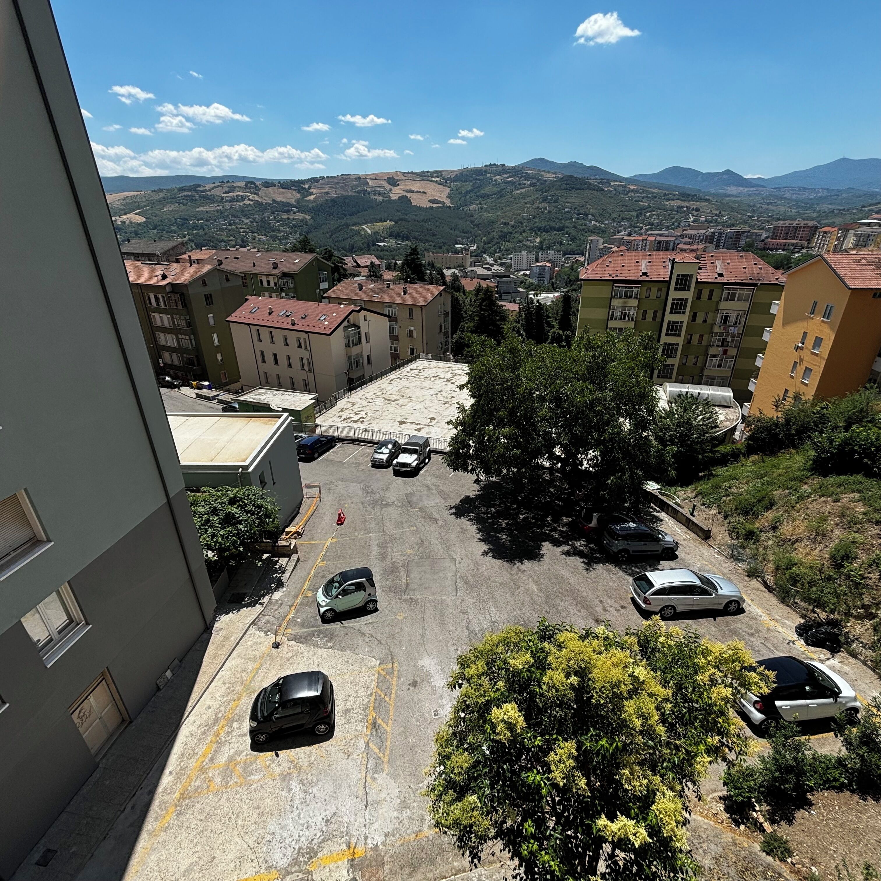 A parking area with several cars, surrounded by buildings with red-tiled roofs and a mountainous landscape in the background.