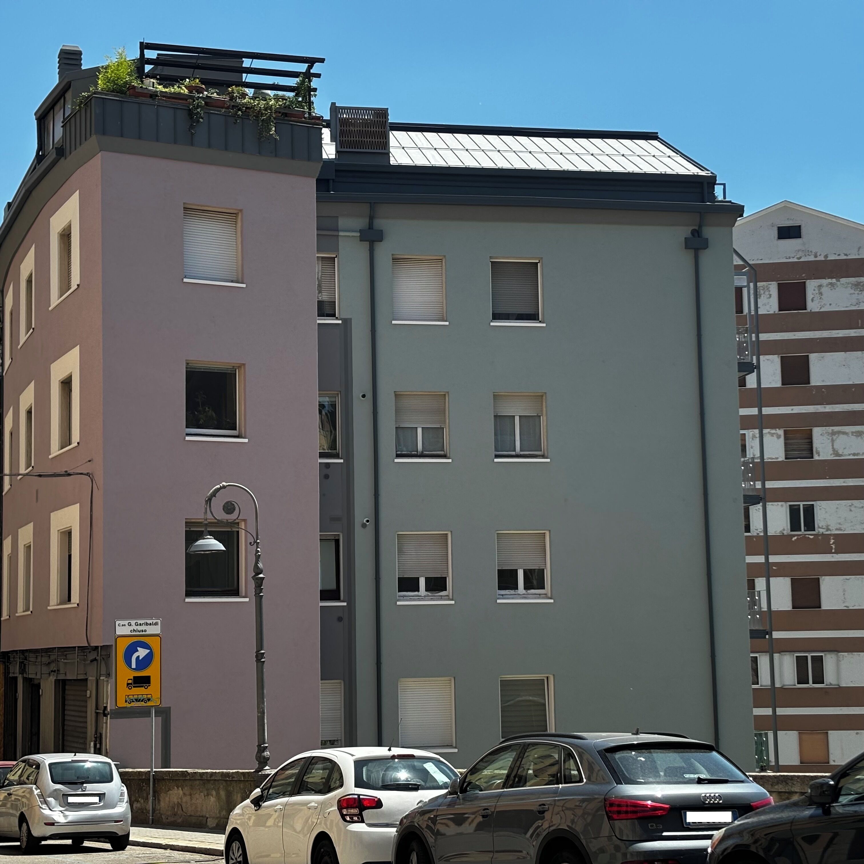 A multi-story building with a mix of residential and commercial spaces. There are several parked cars along the street, and a traffic sign indicating a right turn ahead.
