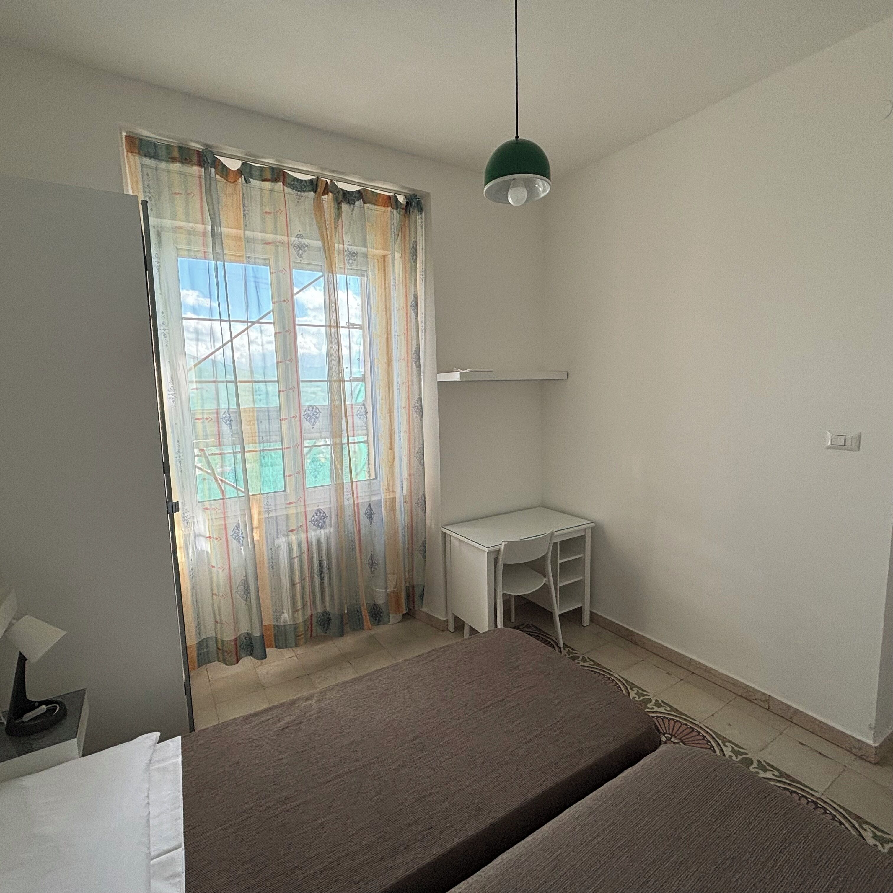 A room with a bed, a desk, and a chair. There is a window with curtains, and a hanging light fixture.