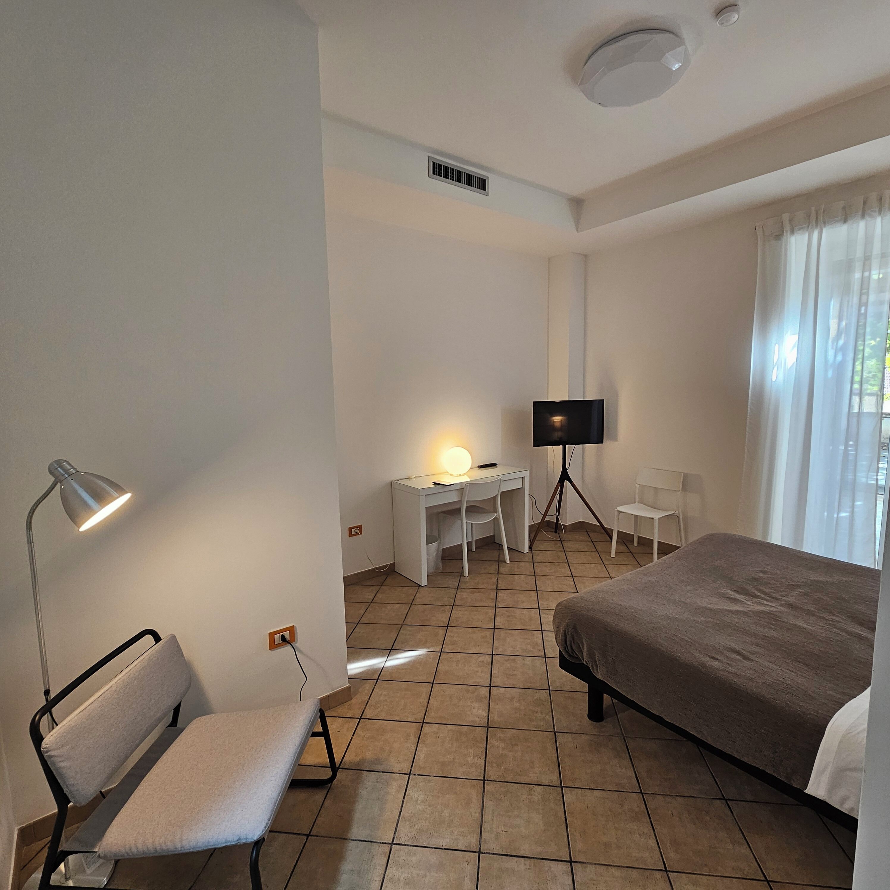 A hotel room with a bed, a desk with a chair, a TV on a tripod, a lamp, and a window with curtains.