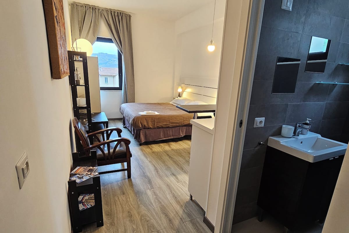 Ein kleines Hotelzimmer mit Bett, Schreibtisch und Stuhl, einem kleinen Tisch mit Zeitschriften, einem Fenster mit Vorhängen, einem Badezimmer mit Waschbecken und Spiegel sowie Blick nach draußen.