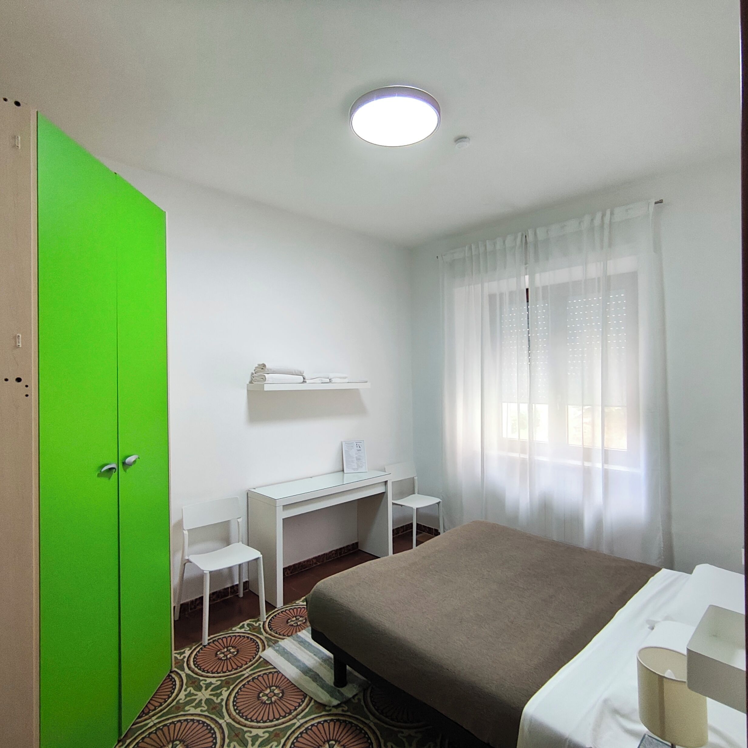 A hotel room with a bed, a desk, a chair, a green wardrobe, and a window with sheer curtains.