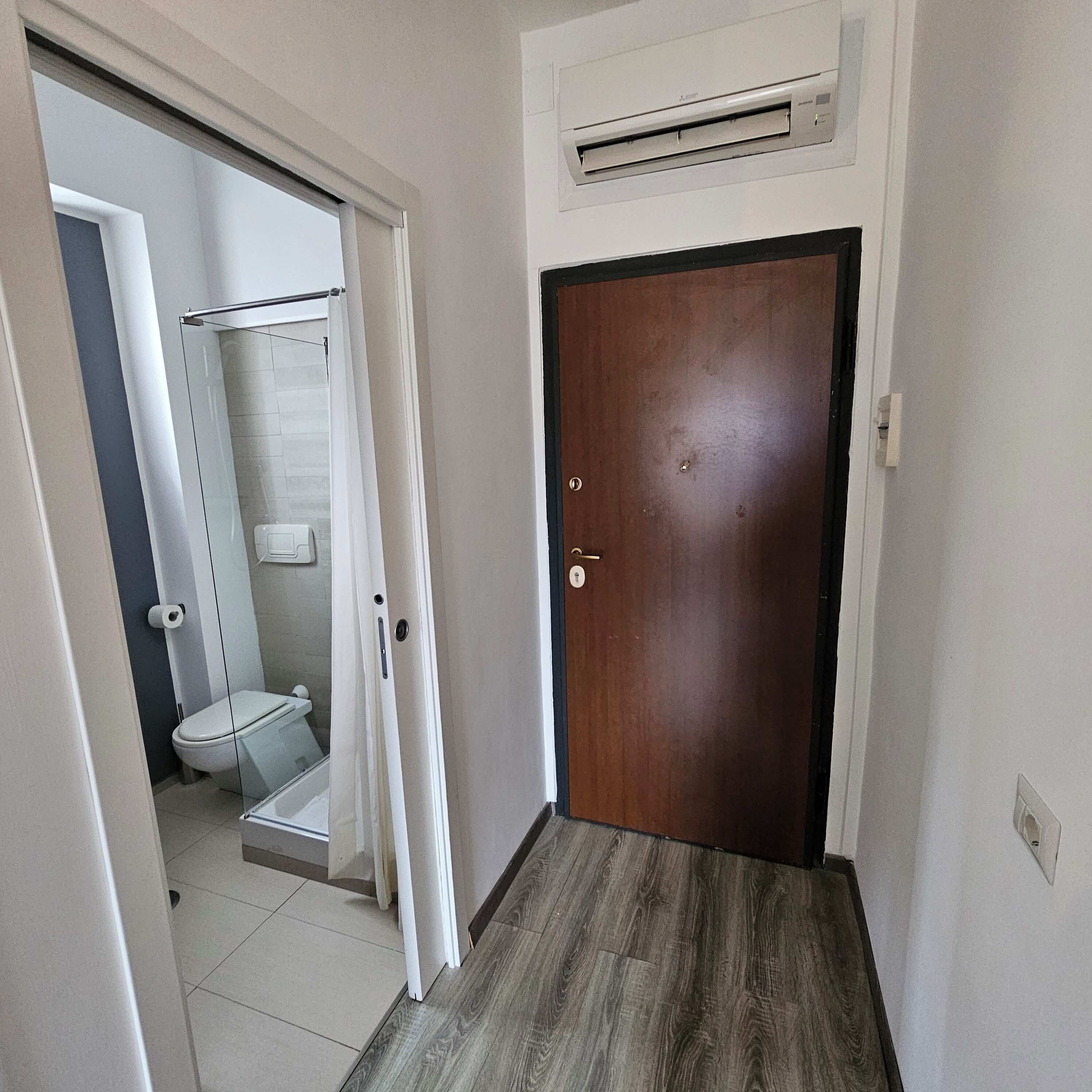 A hallway leading to a bathroom with a toilet, shower, and an air conditioning unit above the door.