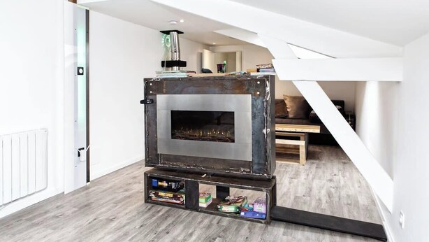A compact, rustic wood-burning stove in a modern loft bedroom with a sloped ceiling and a view of the living area.