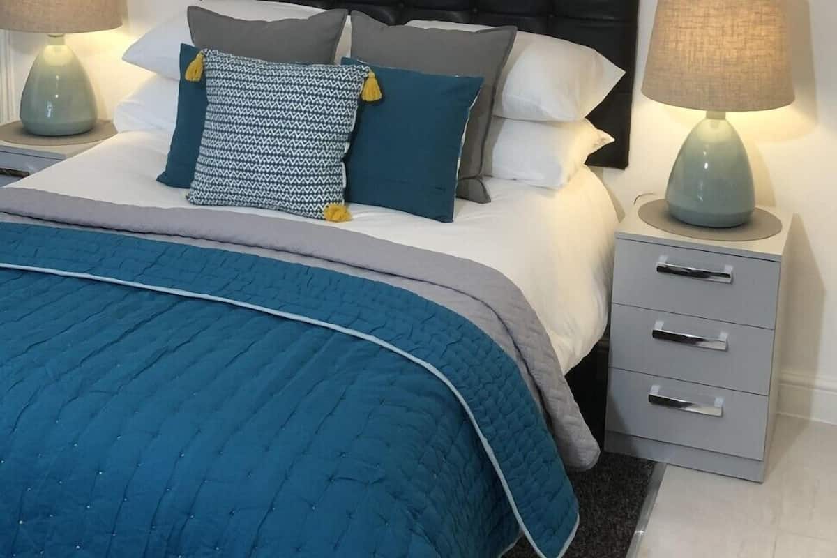 A neatly arranged bedroom with a black tufted headboard, a blue quilted bedspread, and a grey bedside table with a lamp.