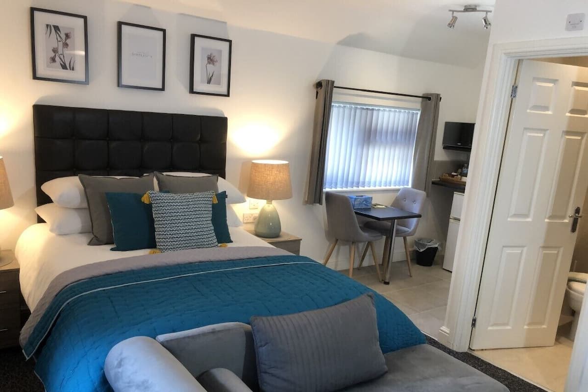A hotel room with a large bed, a desk, and a chair. There are framed pictures on the wall, a lamp, and a window with blinds.