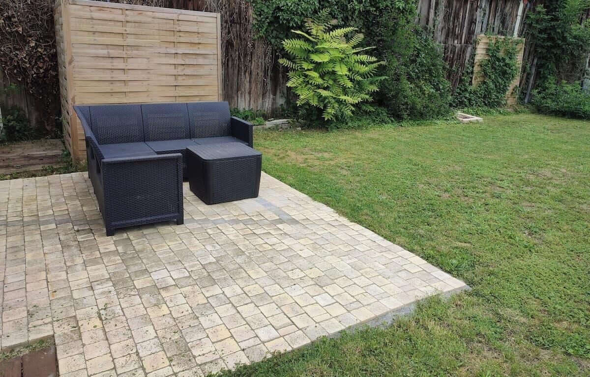 A patio area with a dark-colored sofa and matching ottoman, surrounded by a brick floor and a wooden fence.