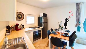 Eine kompakte Küche mit Spüle, Herd und Kühlschrank. Ein Esstisch mit Stühlen steht in der Nähe eines Fensters mit Vorhängen.