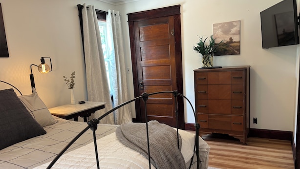 Een slaapkamer met een bed, nachtkastje, lamp, gordijnen, een houten deur, een dressoir en een televisie aan de muur.