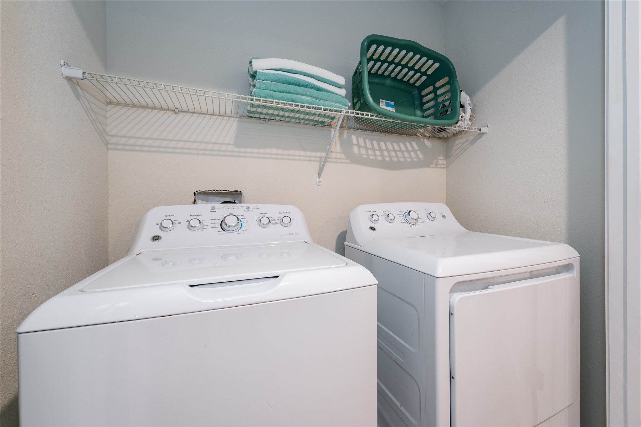 Un espace buanderie comprenant deux machines à laver à chargement frontal blanches et une étagère en métal au-dessus, où sont pliées des serviettes et où se trouve un panier à linge vert.