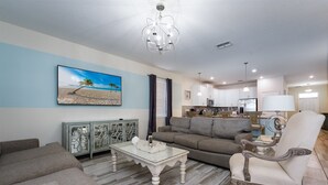 A living room with a sofa, armchair, and coffee table. There is a painting on the wall, a chandelier, and a kitchen in the background. - 9BR Villa Home Theater Pool Sleeps 16 (Davenport)