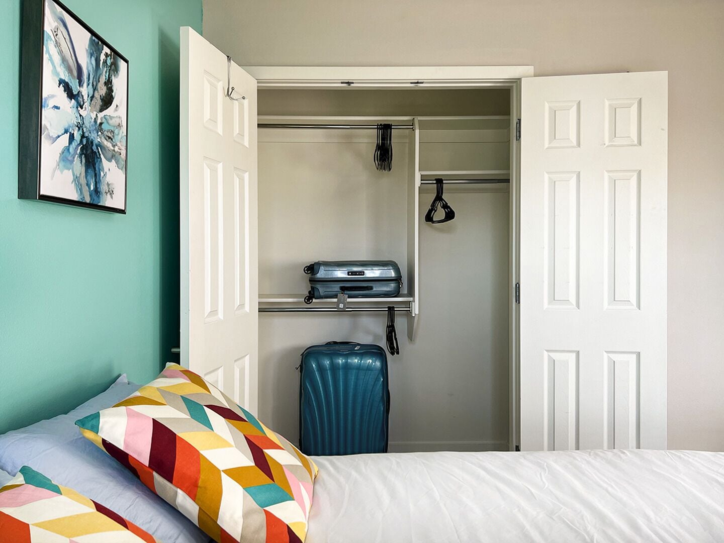 Un dormitorio con cama, almohadas coloridas, una maleta y un armario con ropa colgada.