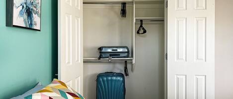 Un dormitorio con cama, almohadas coloridas, una maleta y un armario con ropa colgada.