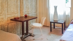 A room with a bed, a small table with a red object, a white chair, and a white vase on a small table.