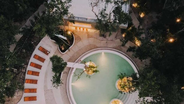 An aerial view of a resort with a circular pool surrounded by a white deck, benches, and a small building with lights.