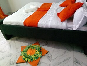 A bed with white and orange bedding, a dark-colored headboard, and a decorative orange and green arrangement on the floor.