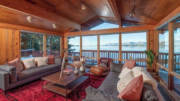 A spacious living room with a large window overlooking a lake and mountains, wooden ceiling, and a cozy seating area with a coffee table and cushions. - Paradise cove - A Mid-Century Modern Waterfront Retreat (Ketchikan)