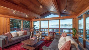 A spacious living room with a large window overlooking a lake and mountains, wooden ceiling, and a cozy seating area with a coffee table and cushions.