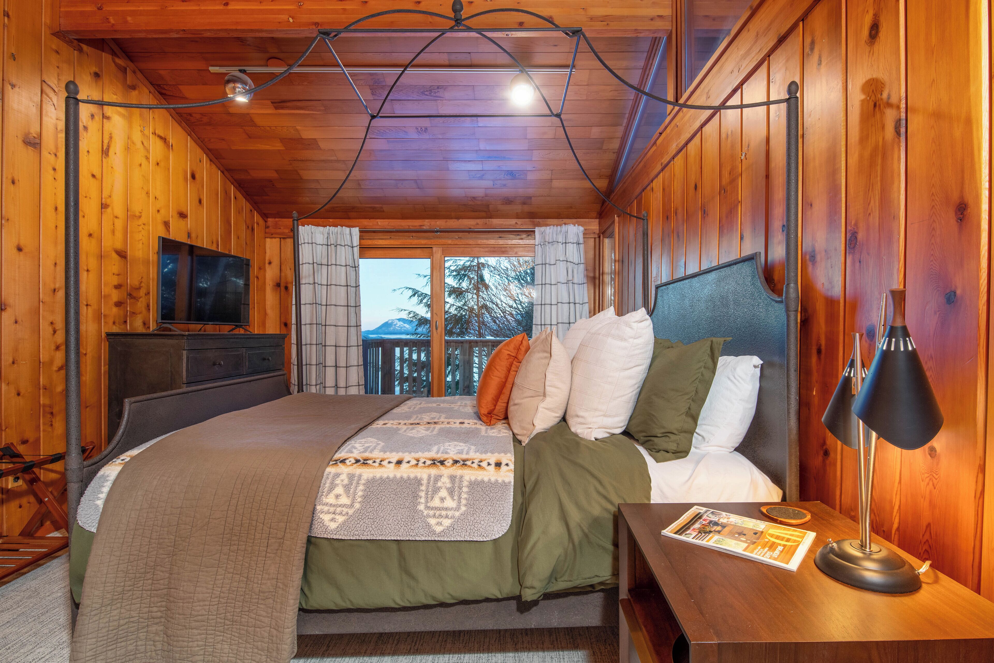 Une chambre à coucher dans une cabane en bois, avec un lit, une table de chevet, une lampe, un téléviseur et une vue sur l’extérieur.