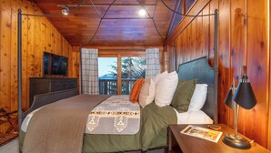 A wooden cabin bedroom with a bed, a nightstand, a lamp, a TV, and a view of the outdoors. - Paradise cove - A Mid-Century Modern Waterfront Retreat (Ketchikan)