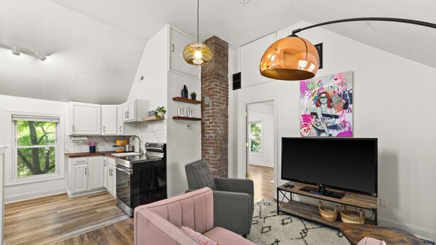 A modern kitchen with white cabinets, a brick accent wall, and a dining area with a pink sofa and a TV. - Cozy Loft Apartment in Ohio City (Cleveland)